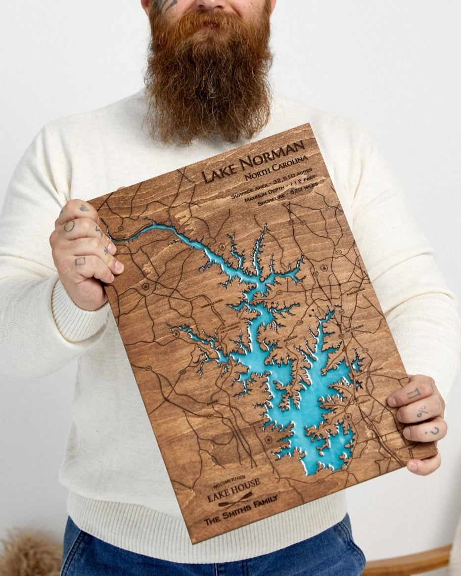 Man holding a personalized Lake Norman wood and blue epoxy map with engraved roads and a family lake house inscription.