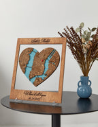Heart-shaped wooden map with engraved text on a table with a vase and flowers.