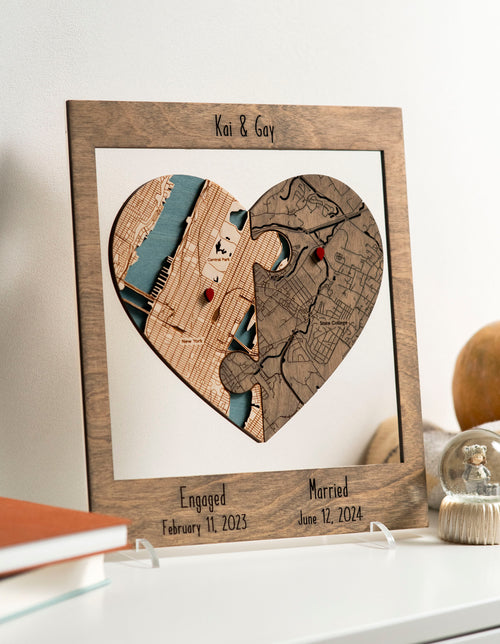 Customized heart-shaped map puzzle in a frame by a person against a wall, featuring two locations with dates for engagement and married