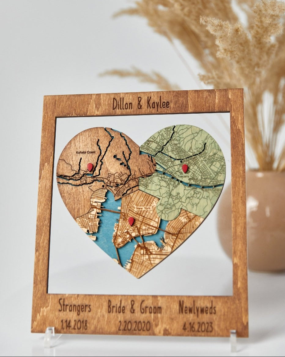 Heart-shaped map cutout with personalized text on a wooden frame, displayed against a neutral background.