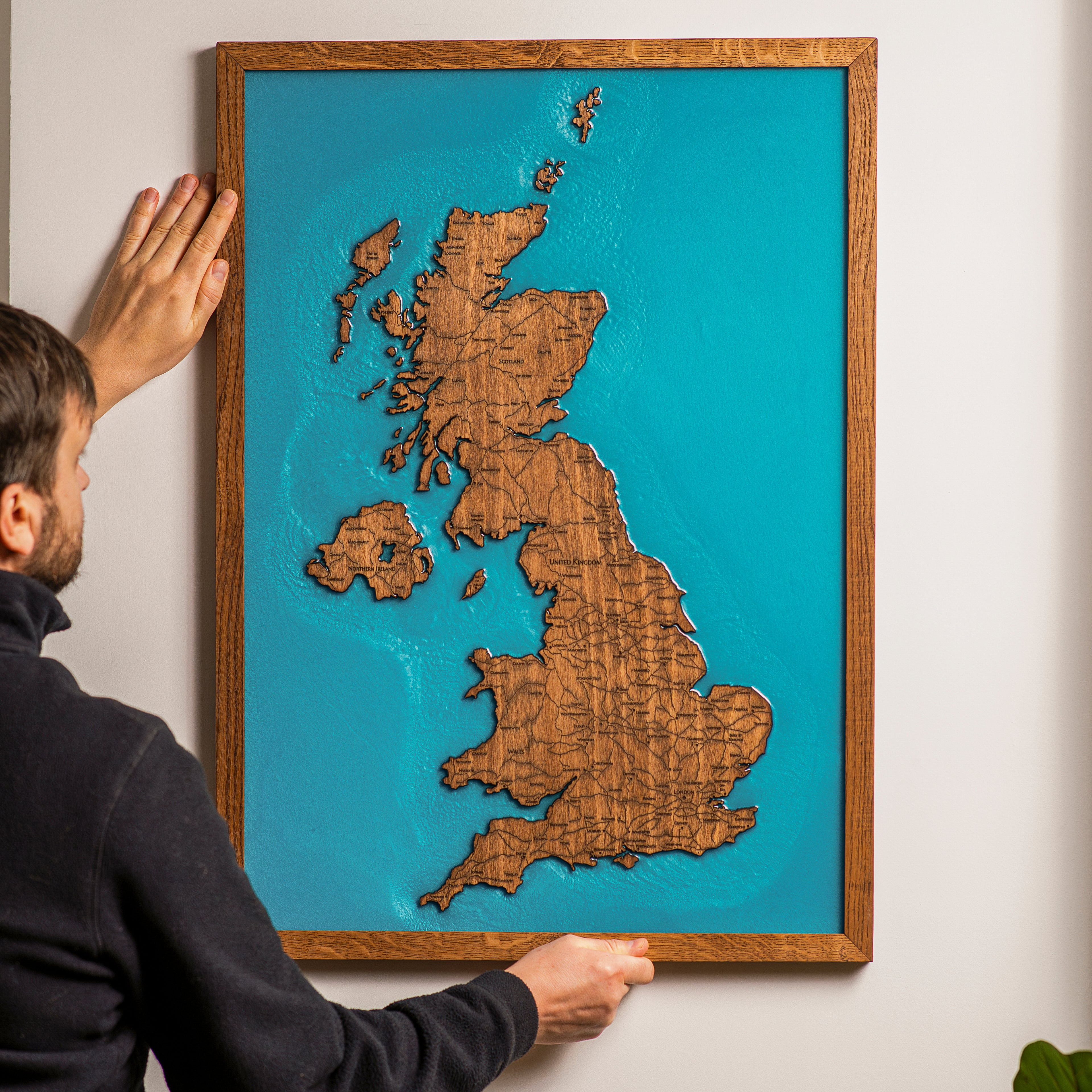 Person hanging framed United Kingdom wooden map on wall showing easy installation and real scale