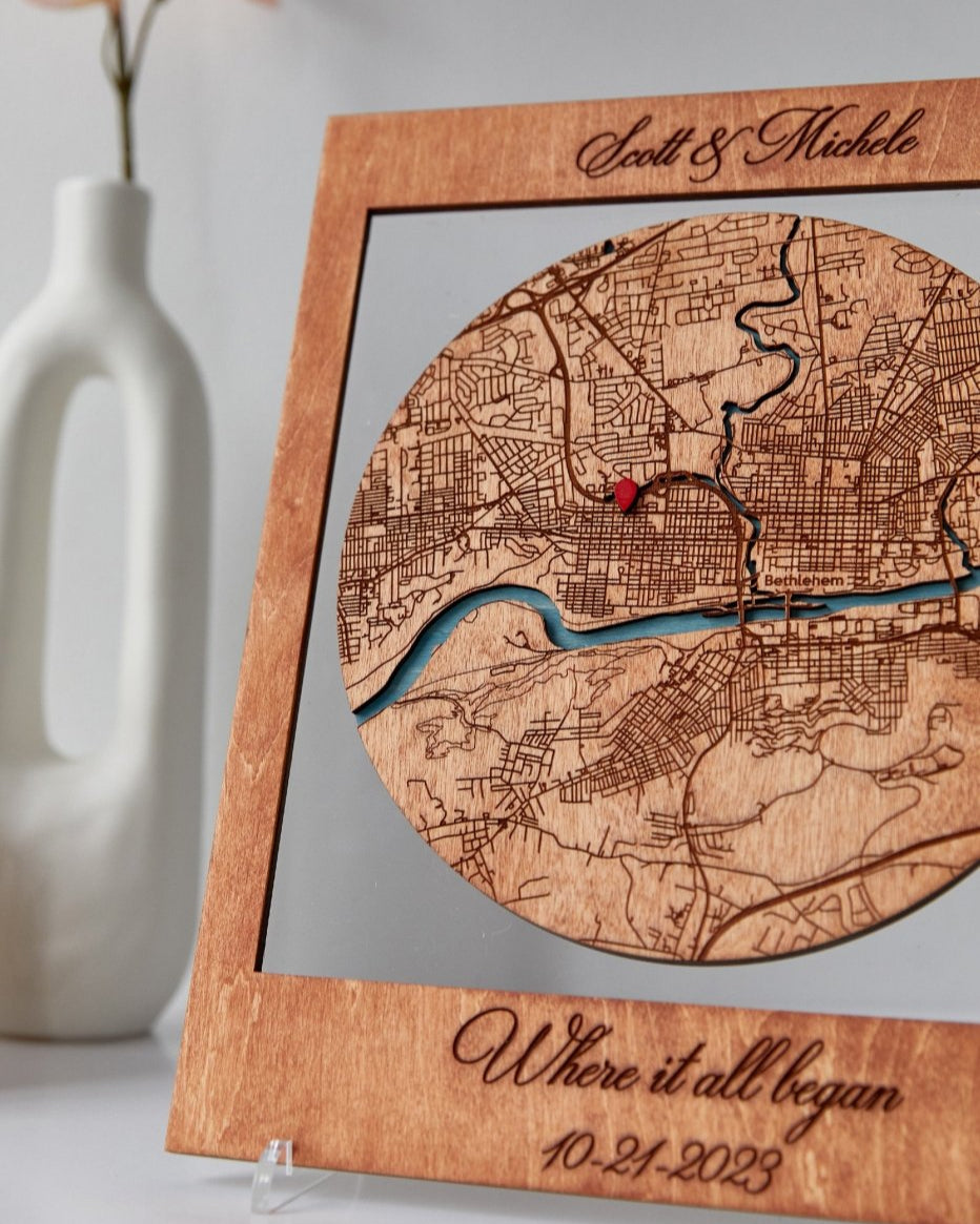 Wooden map with engraved text and a red dot, placed on a stand with a vase in the background.