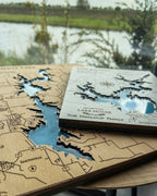 Wooden map with laser-cut details of a lake house and a book titled 'Lake House' by the Maalouf Family on a wooden surface with a lake view.