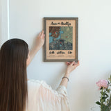 Woman holding a Classic Square three-location wooden map in a Grey oak frame against a wall.