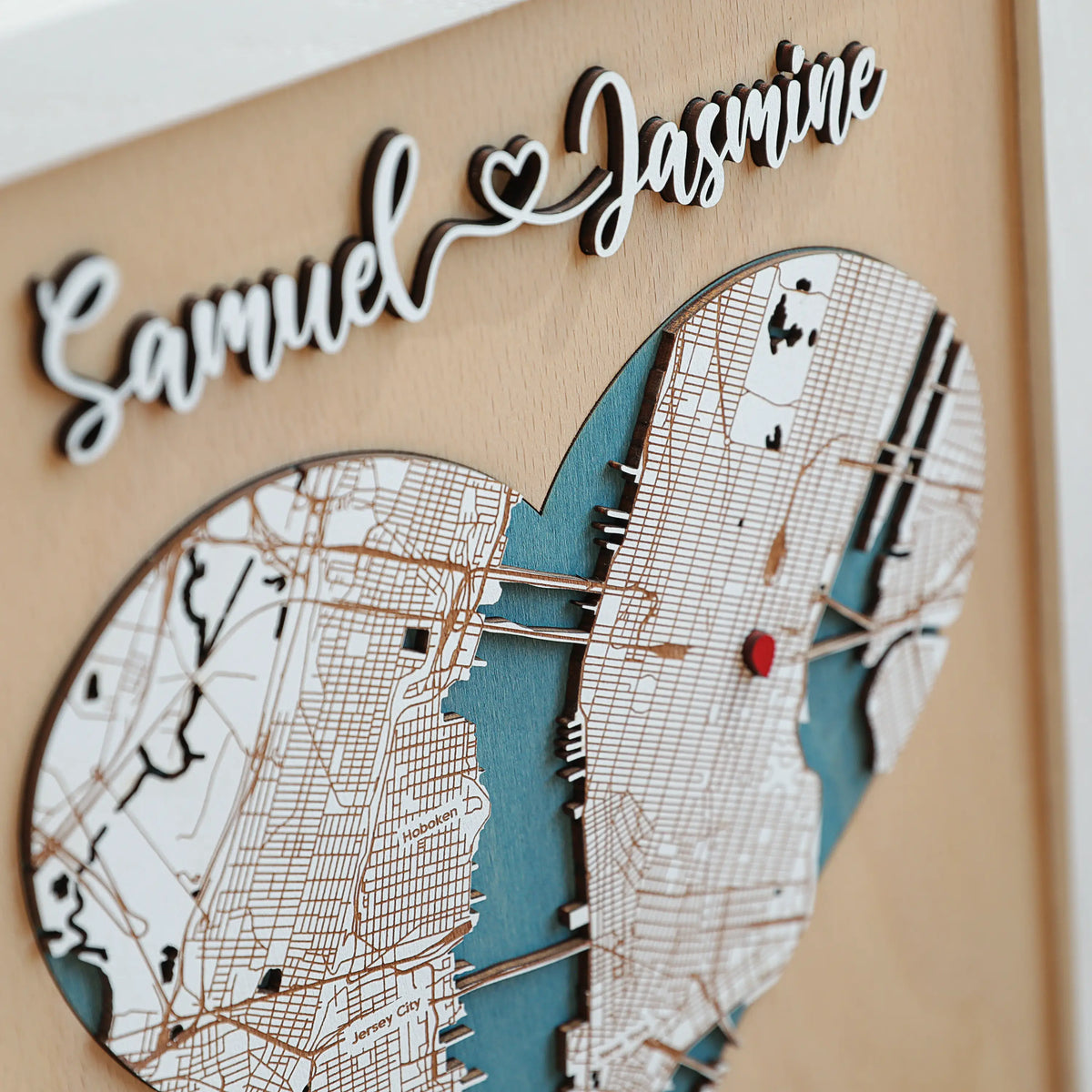 Close-up of personalized 3D wooden heart map showing New York location details, layered white map pieces, blue water inlay, red marker, and raised names.