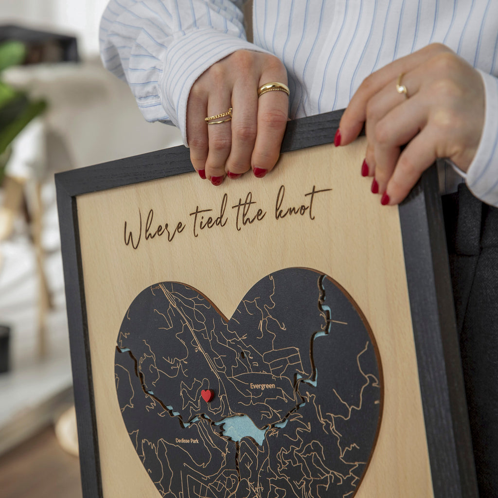 Close-up of black heart-shaped wooden map with red pin, custom location details, and wedding phrase in oak frame.