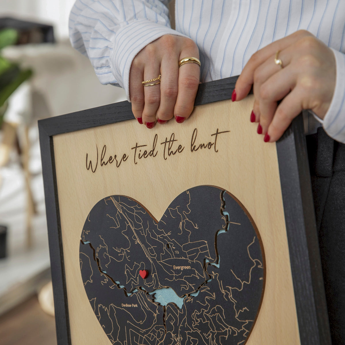 Close-up of black heart-shaped wooden map with red pin, custom location details, and wedding phrase in oak frame.