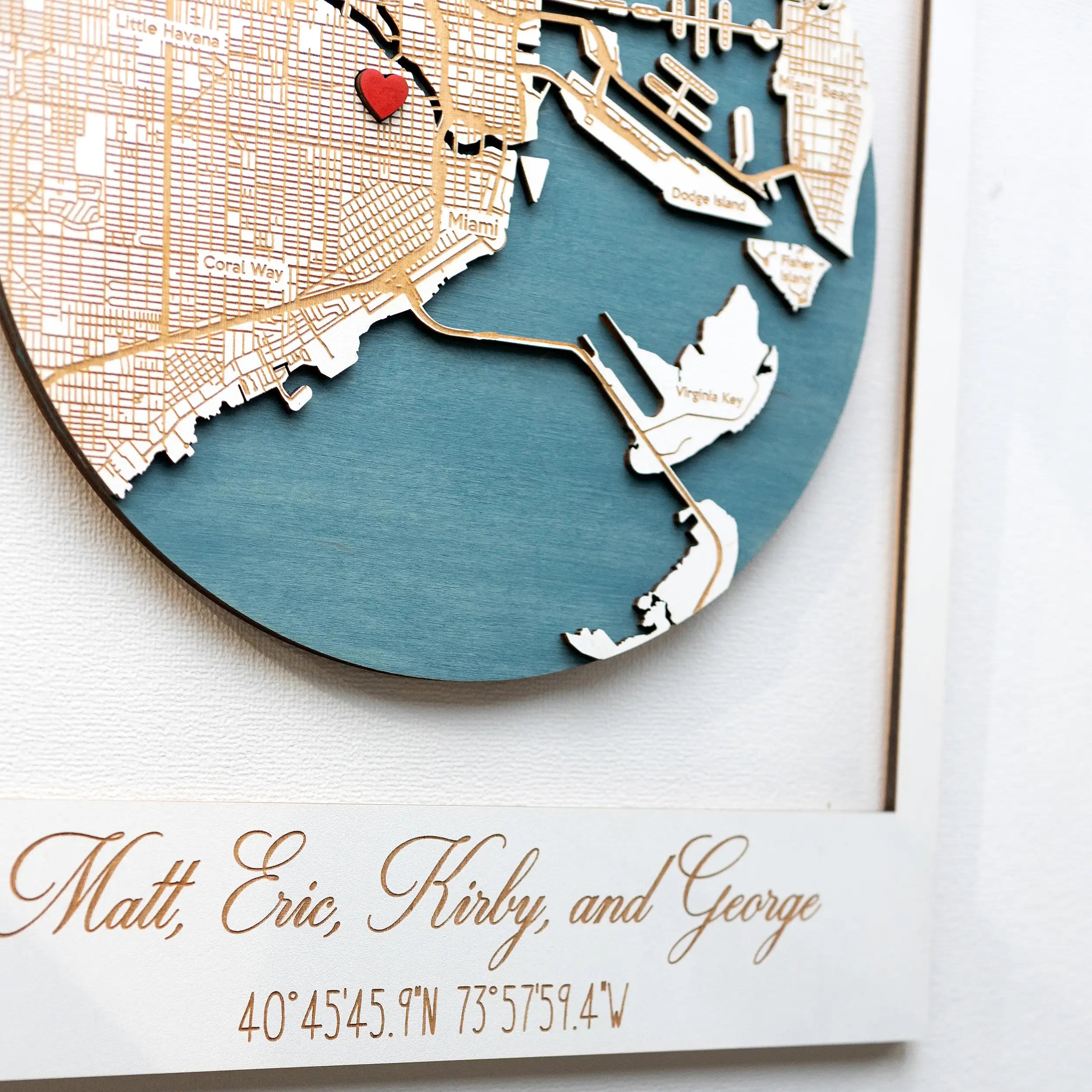 Close-up of a personalized 3D wooden Miami map inside a white frame, showing layered coastline details, blue water, engraved roads, and custom family name text.