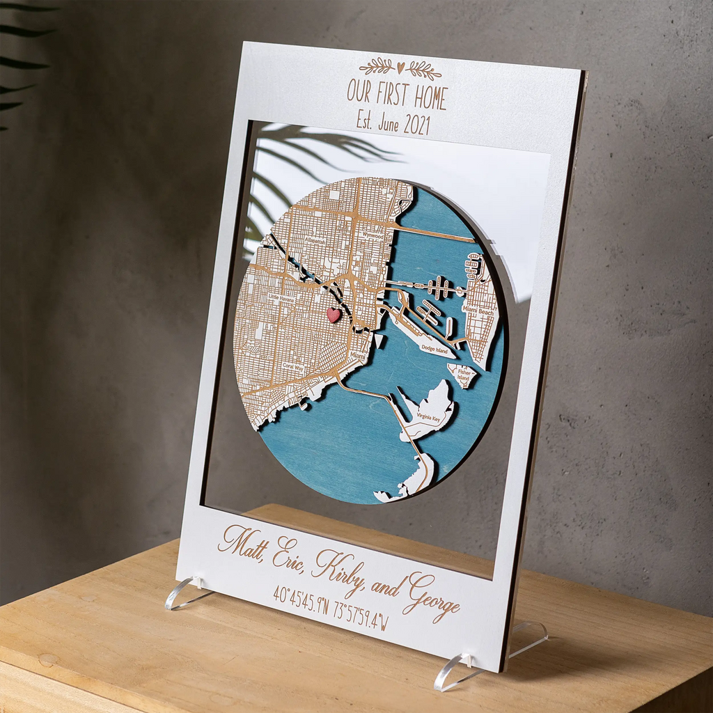 White framed personalized wooden location art displayed on a tabletop stand, custom housewarming gift with 3D Miami map, heart pin, and family names.