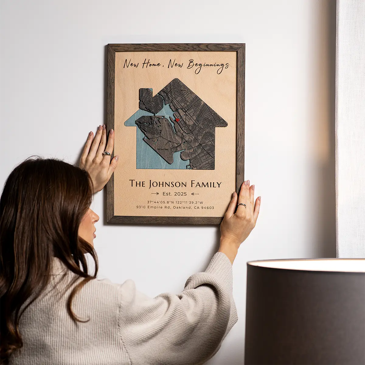 Person hanging a house-shaped personalized wood map frame on the wall with “New Home, New Beginnings” and family name.