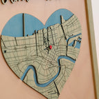 Close-up of heart-shaped 3D wooden New Orleans location map with blue river details, engraved streets, red location marker, and layered wood design.
