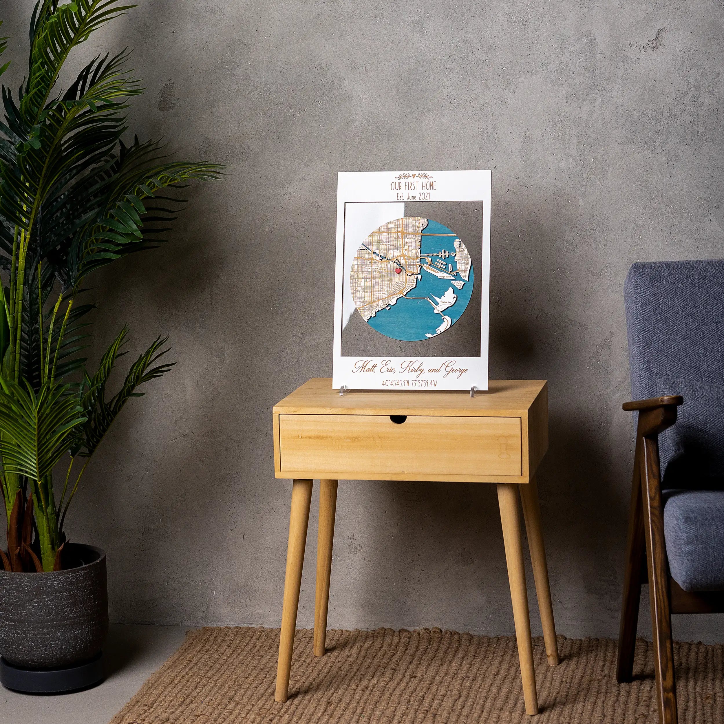 White framed custom housewarming wall art displayed on a wooden side table, featuring a layered Miami wooden map with blue water details and heart pin.