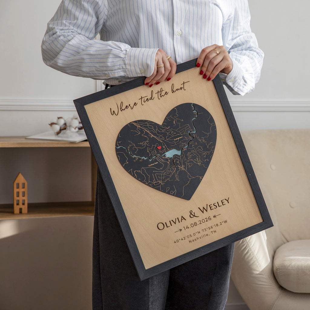 Person holding a framed heart-shaped map with location where it all began and personalized text in a room.