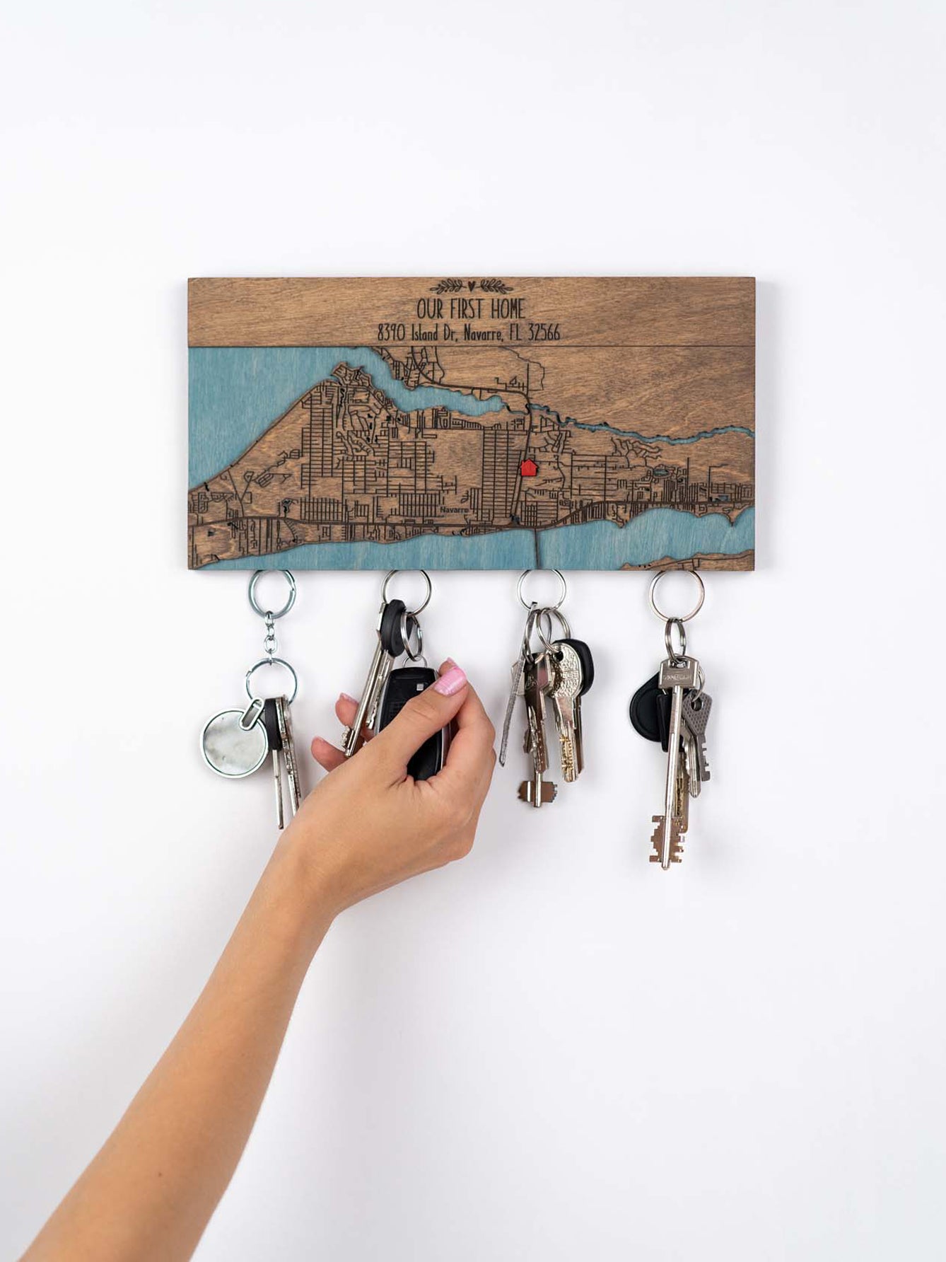 A person is hang  a set of keys on a magnetic wooden key holder with an engraved custom location map and text that reads "OUR FIRST HOME"