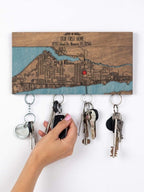 A person is hang  a set of keys on a magnetic wooden key holder with an engraved custom location map and text that reads "OUR FIRST HOME"