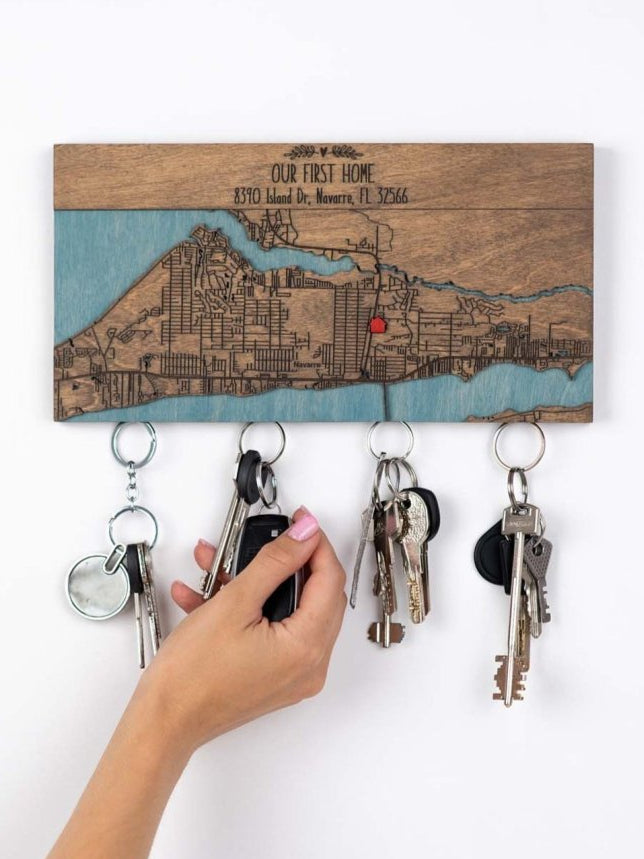 A person is hang  a set of keys on a magnetic wooden key holder with an engraved custom location map and text that reads "OUR FIRST HOME"