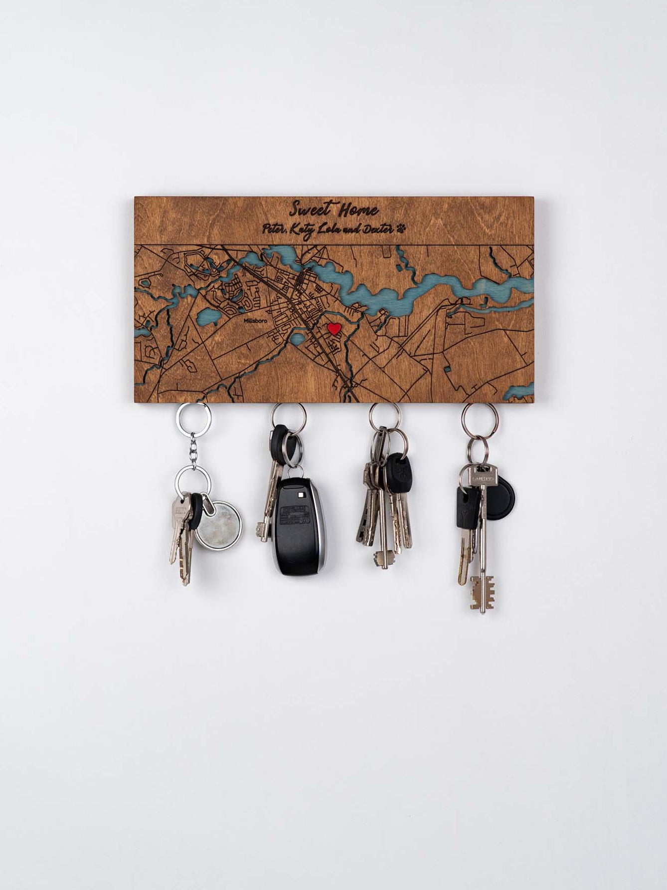 Wooden key holder mounted on the wall. The key holder is engraved with a map design and the phrase "Sweet Home," personalized with additional names of the owners of the house. the decorative map, there are strong magnetics from which four sets of keys are hanging, including one set with a car key fob. The is a plain, deep brown, colored surface, emphasizing the aesthetic and functionality of the magnetic wooden key holder.