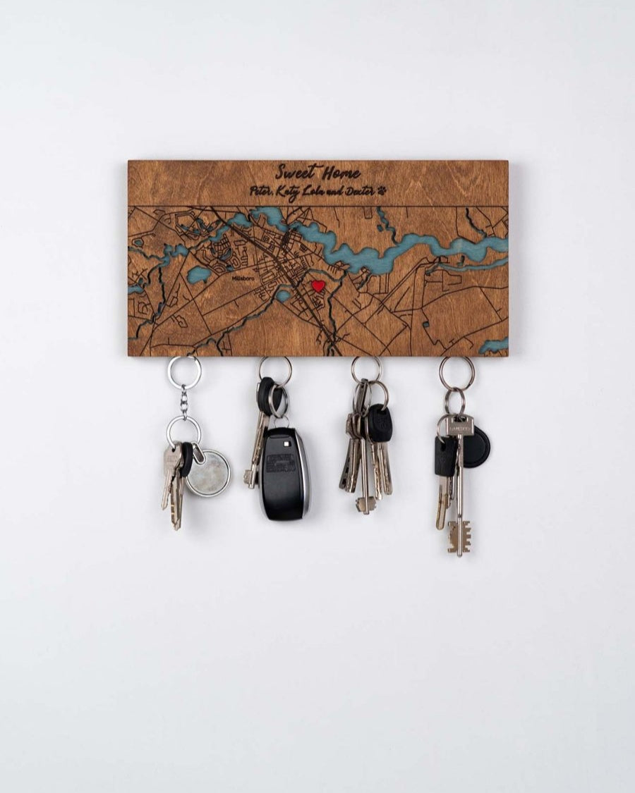 Wooden key holder mounted on the wall. The key holder is engraved with a map design and the phrase "Sweet Home," personalized with additional names of the owners of the house. the decorative map, there are strong magnetics from which four sets of keys are hanging, including one set with a car key fob. The is a plain, deep brown, colored surface, emphasizing the aesthetic and functionality of the magnetic wooden key holder.