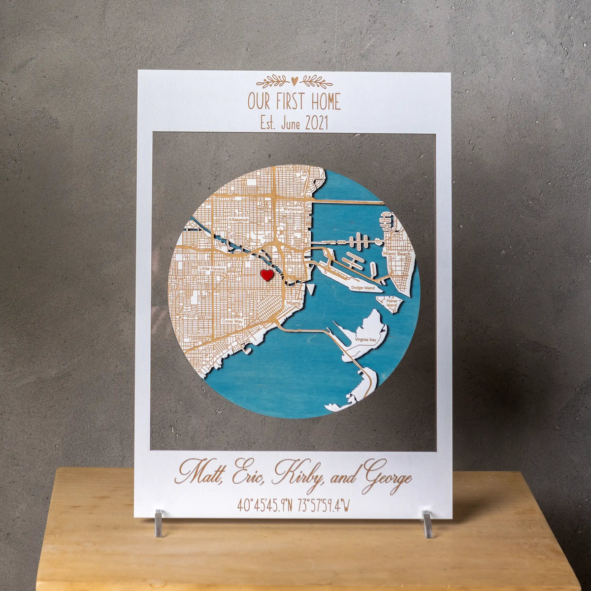 Front view of a custom 3D wooden location map in a white frame with Our First Home text, Miami map design, heart marker, and personalized family names.