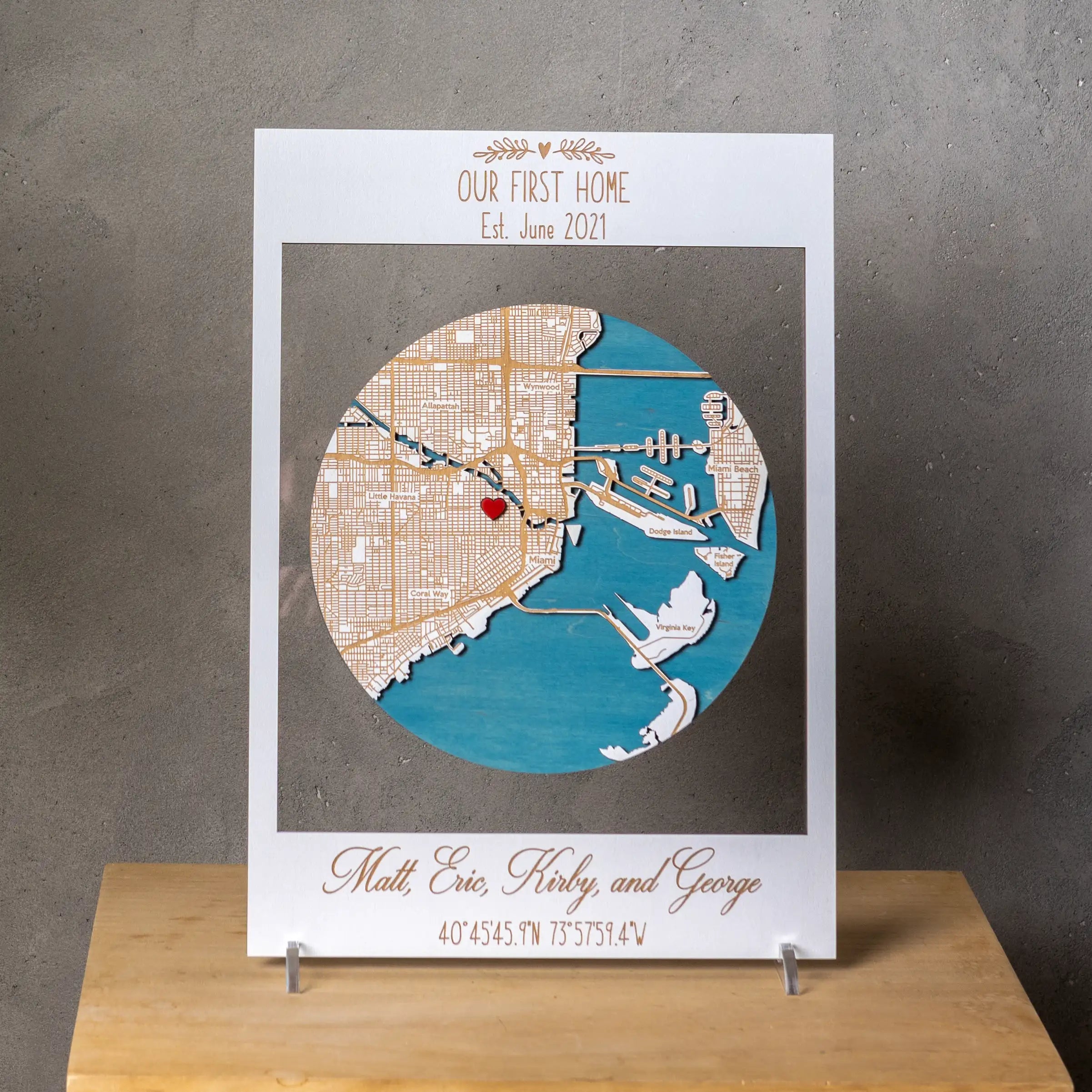 Front view of a custom 3D wooden location map in a white frame with Our First Home text, Miami map design, heart marker, and personalized family names.