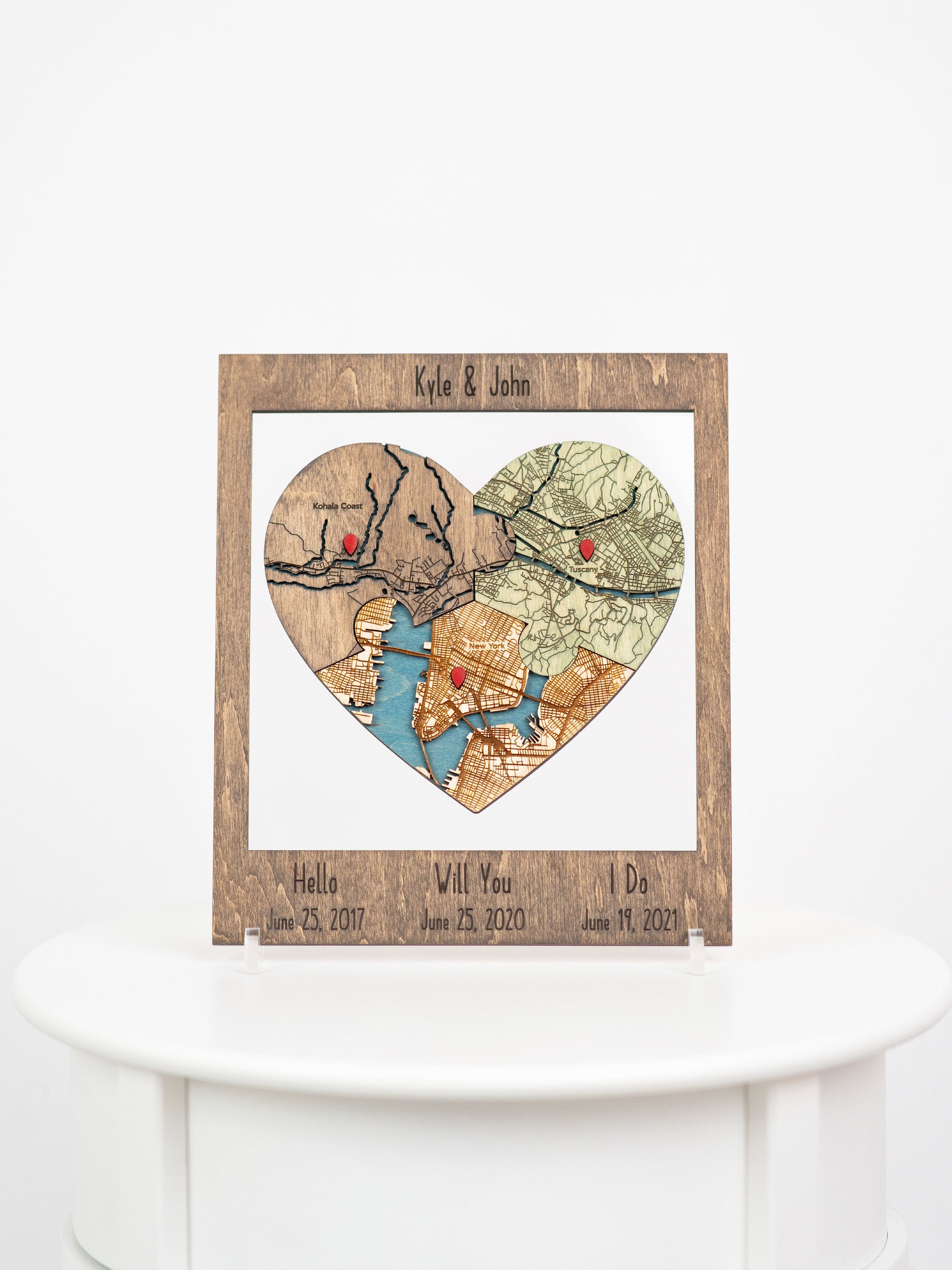 Personalized wooden frame with heart shaped cutout displaying three segments of maps, each marked with a red pin. The custom names are engraved at the top. Below the maps, there are engraved statements and dates: "Hello "Will You and "I Do. Commemorates significant milestones in couples' relationships on those dates - their meeting, engagements, and marriages. This custom piece serves as an anniversary gift that tells the story of their journey together using map locations significant to their relationship.