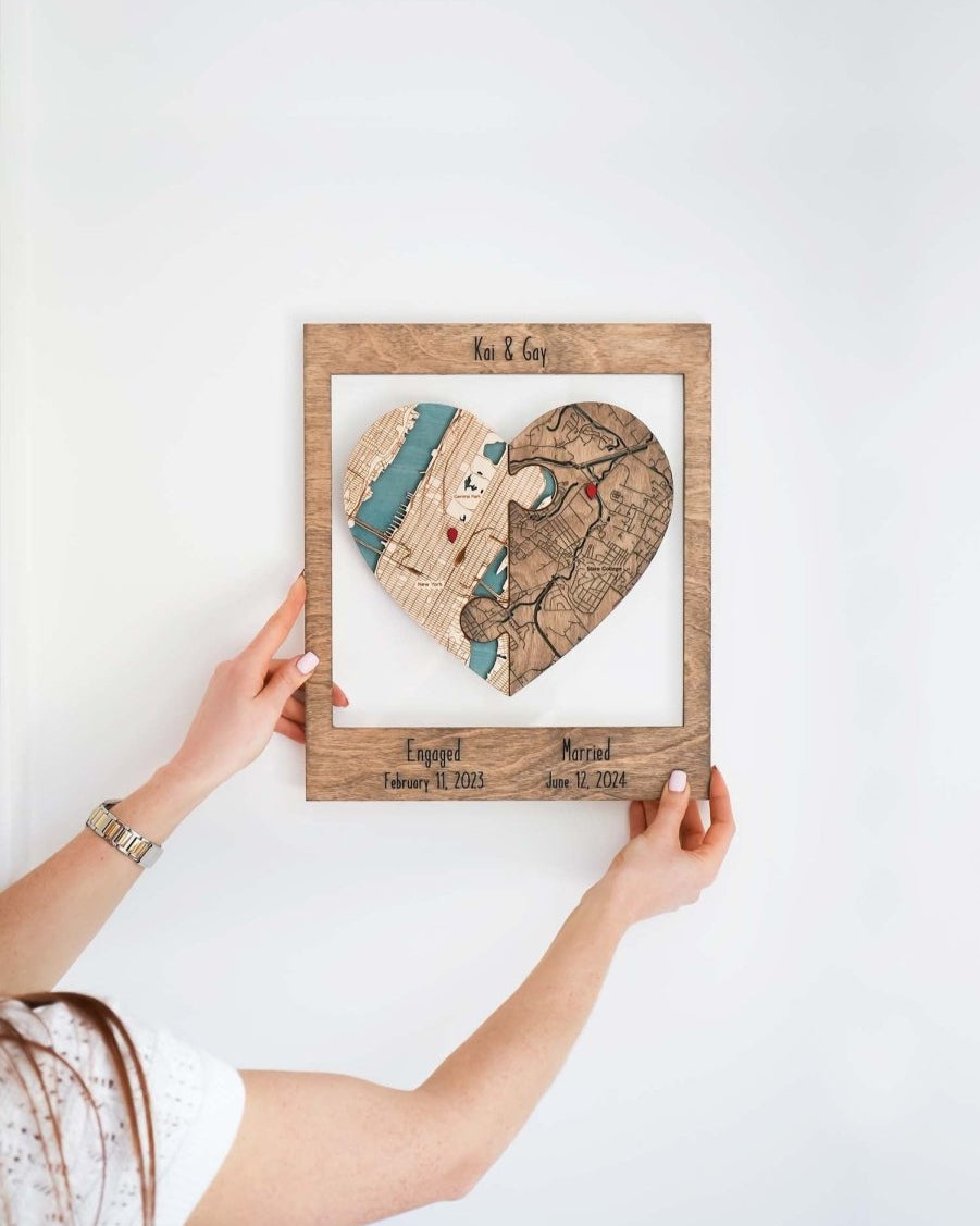 Customized heart-shaped map puzzle in a frame by a person against a wall, featuring two locations with dates for engagement and marriage
