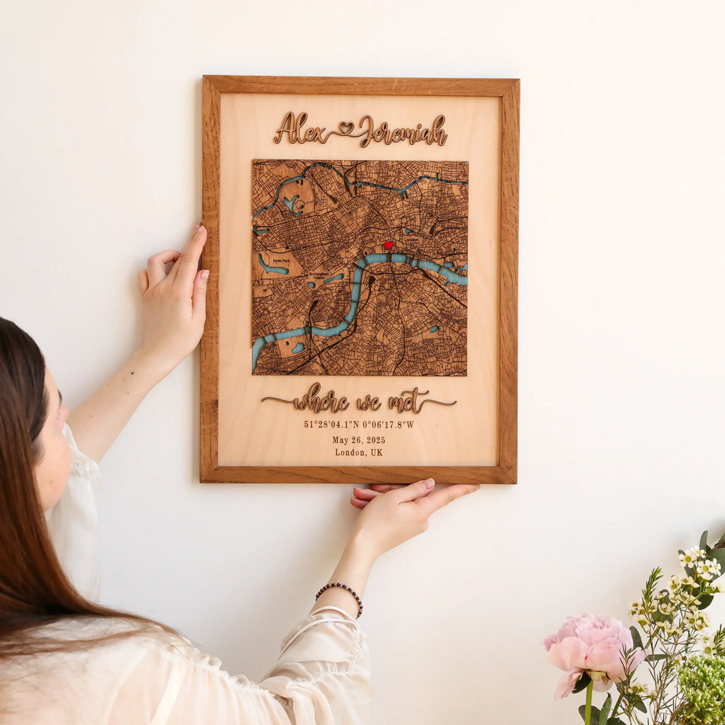 Person hanging a personalized 3D wooden map on the wall, showing custom names, London location map, raised lettering, and oak frame.