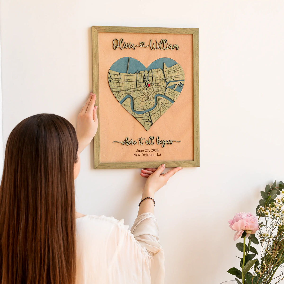 Person hanging a romantic personalized anniversary map on the wall, featuring a heart-shaped New Orleans location map, raised names, custom date, and sage frame.