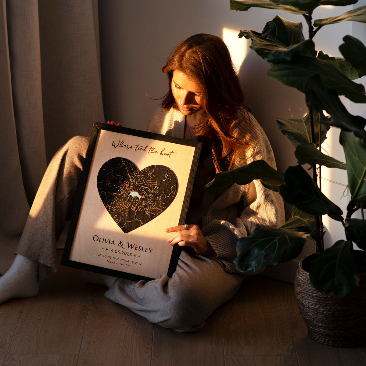 Person holding a framed heart-shaped map with 'Olivia & Wesley' and a date, sitting on the floor with a plant.