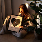 Person holding a framed heart-shaped map with 'Olivia & Wesley' and a date, sitting on the floor with a plant.