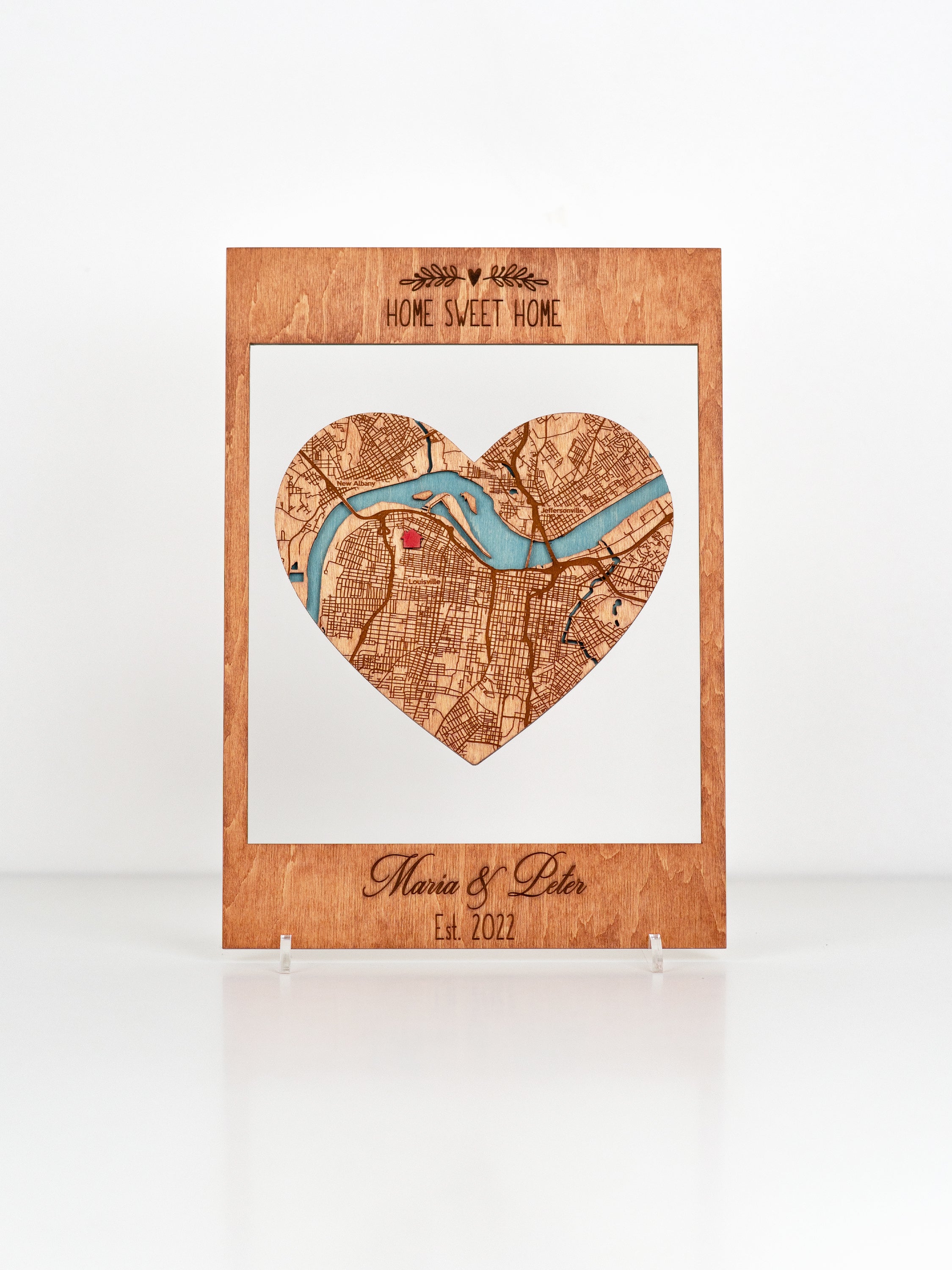 A wooden plaque with a heart-shaped cutout featuring a map design, inscribed with "Home Sweet Home" at the top and "Maria & Peter Est. 2022"  and You can add any text.